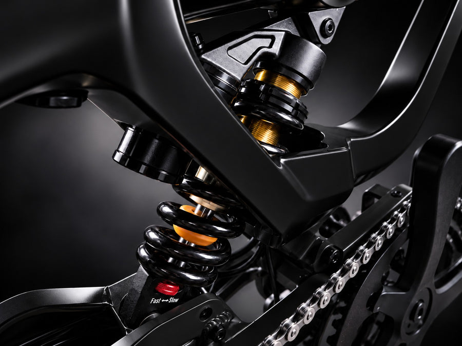 Close-up of a black motorcycle rear suspension system featuring a black coil spring with gold-colored shock absorber elements, set against a dark studio background.
